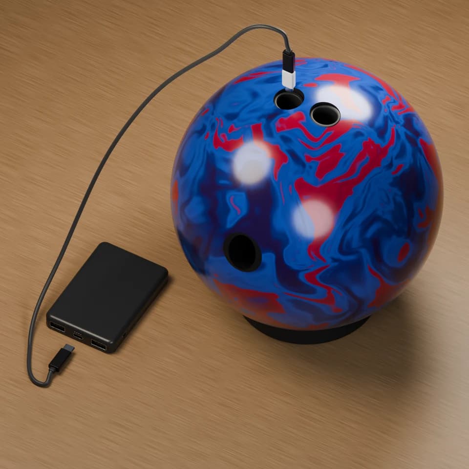 A bowling ball rests on a ball cup beside the power bank. The white adapter is shown above the middle finger hole, ready to lower through the grip to the installed sensor. Connect the black end to the center USB-C port on the power bank. The grip stays installed.